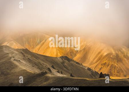 Blick auf goldfarbene Berge, die in ätherischen Nebel gehüllt sind, einen zerklüfteten Pfad, der sich durch die vulkanische Landschaft schlängelt, eine Symphonie aus Erdtönen, Landmannal Stockfoto