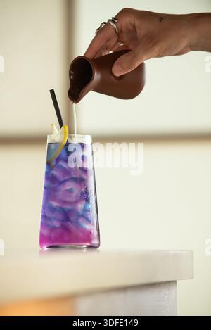 Gießen Sie Flüssigkeit von Hand in ein violettes Eisgetränk in einem klaren Glas mit Zitronengarnitur und Stroh, fotografiert auf einem sauberen Studiohintergrund. Stockfoto