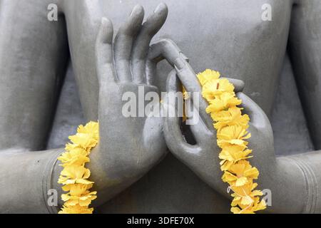 Das Detail der Buddha-Statue in Meditationsposition. Hände, die Buddha mit einem Kranz aus gelben Blumen meditieren Stockfoto