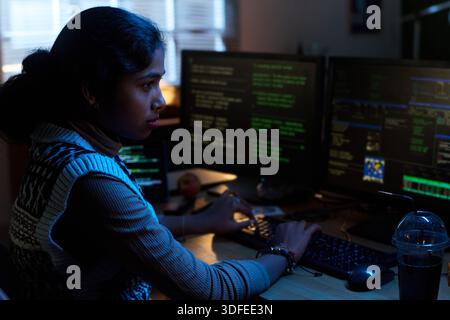 Junge südasiatische Frau, die an Computermonitoren in Dark Office arbeitet Stockfoto