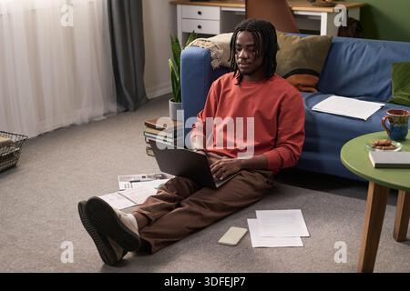 Junger schwarzer Mann, der auf dem Boden sitzt und einen Laptop für Fernstudien benutzt Stockfoto