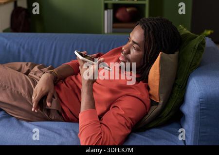Junger schwarzer Mann, der sich auf dem Sofa mit dem Smartphone-Sprachassistenten entspannt Stockfoto