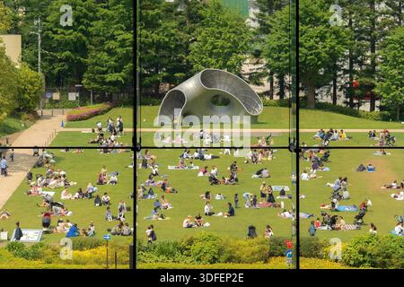Blick auf die Menschen, die sich auf einem lebendigen grünen Rasen in der Innenstadt von Tokio unter dem warmen Sonnenlicht entspannen, durch ein Fenster mit einer modernen Skulptur Stockfoto