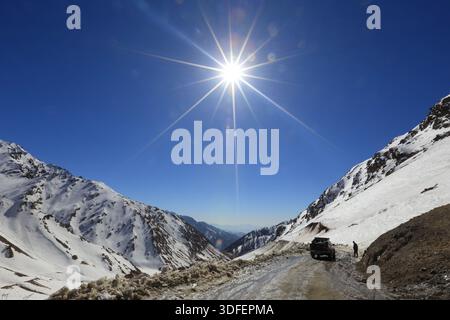 Blick auf die Sonne, die hell über den schneebedeckten Bergen scheint, ein Fahrzeug und eine Person auf der Straße im Tal, Lowari Pass, Khyber Pakhtunkhwa, Pa Stockfoto