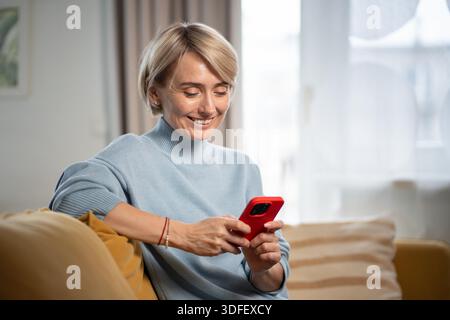 Glückliche, begeisterte Frau mittleren Alters, die Nachrichten schickt, online mit Freunden auf dem Smartphone chatten, auf dem Bildschirm lächelt, Text eingibt, Medien-Feeds durchblättert. Positive Frauen, die freie Zeit beim Telefonieren verbringen. || Modell freigegeben Stockfoto