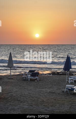 Der Blick auf die warme Sonne taucht tief und strahlt einen goldenen Glanz auf dem ruhigen Strand mit Sonnenschirmen und Stühlen mit Blick auf das Meer, Paphos, Paphos, Zypern. Stockfoto