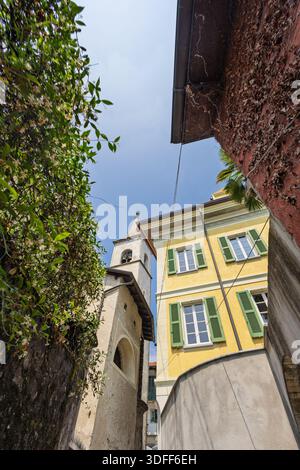 Eine sonnendurchflutete, enge Straße, die von einem gelben Gebäude mit grünen Fensterläden und einer verwitterten Wand eingerahmt wird. Üppiges Grün klettert an den Wänden, während blauer Himmel und ein C Stockfoto