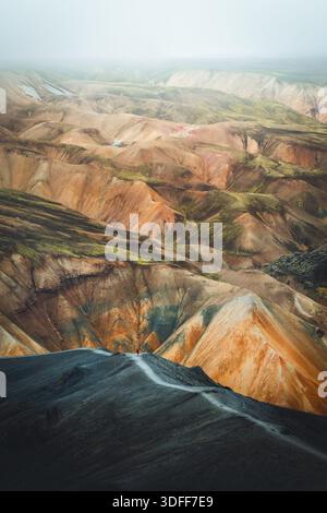 Der Blick auf einen gewundenen Pfad führt durch einen schroffen, dunklen Vorderboden und führt in eine atemberaubende Landschaft mit bunten Hügeln unter einem nebeligen Himmel, Landmannal Stockfoto