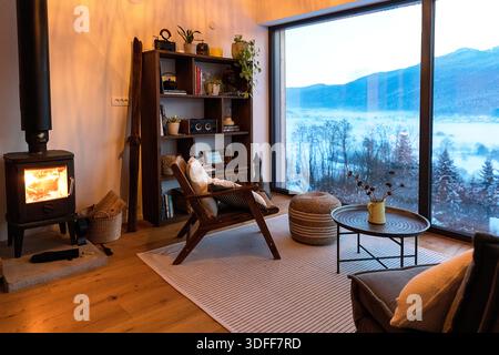 Gemütliches Wohnzimmer im skandinavischen Stil mit Kamin und Panoramafenster, minimalistischen Möbeln, Pflanzen und malerischem Blick auf die Winterlandschaft Stockfoto