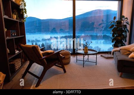 Gemütliches Wohnzimmer im skandinavischen Stil mit Panoramafenster mit minimalistischen Möbeln, Pflanzen und malerischem Blick auf die Winterlandschaft Stockfoto