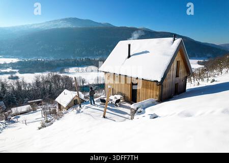 Mann, der auf der Terrasse einer kleinen Holzhütte mit Schnee steht, mit tollem Blick über den Fluss Krka und die Hügel an einem sonnigen Wintertag in Slowenien Stockfoto
