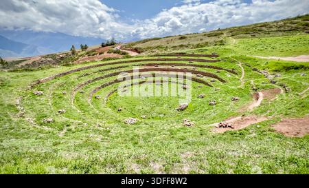 Blick auf konzentrische landwirtschaftliche Terrassen, die in die Erde fallen, ein lebendiges Grün vor dem alten Mauerwerk und dem bewölkten Himmel, Moray, Peru. Stockfoto