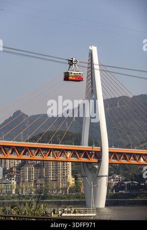 Blick auf eine Seilbahn, die in der Nähe einer weißen Brücke mit orangefarbenen Kabeln über den glitzernden Fluss vor dem Hintergrund der fernen Berge, Chongqing, C, aufsteigt Stockfoto