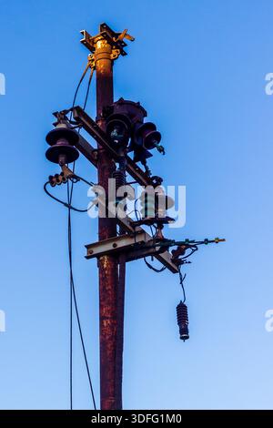 Rostiger Strommast mit Isolatoren gegen klaren blauen Himmel bei Sonnenuntergang Stockfoto