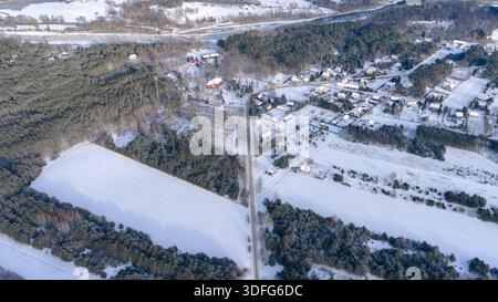 Aus der Vogelperspektive von Nowy Lubiel, Polen, mit einer geraden Straße in Richtung eines Dorfes, Nadelwäldern, Schneefeldern und einem gefrorenen Fluss unter weicher Mittagszeit Stockfoto