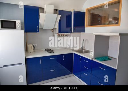 Moderne, kompakte Küche mit blauen Schränken, Waschbecken, Herd und Haushaltsgeräten. Sauberes Appartementdesign mit funktionalem Layout, geeignet für Stockfoto