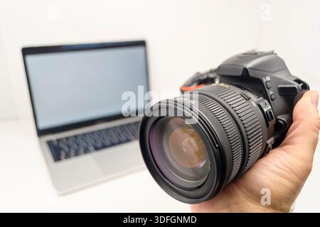 Nahaufnahme einer Hand, die ein DSLR-Kameraobjektiv hält, mit einem unscharfen Laptop im Hintergrund, kreatives digitales Workflow-Konzept. Stockfoto