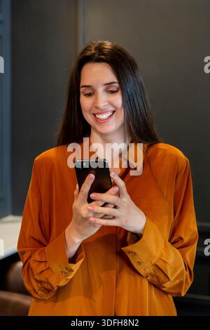 Lächelnde Frau, die ihr Smartphone in einem modernen Innenraum mit dunkler Wand benutzt, lässig stehend und mit einem warmen und freundlichen Ausdruck in A zeigt Stockfoto