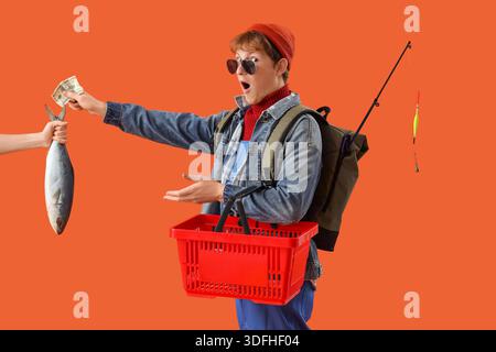 Lustiger junger Fischer mit Warenkorb, der Fisch aus weiblicher Hand auf orangem Hintergrund kauft Stockfoto