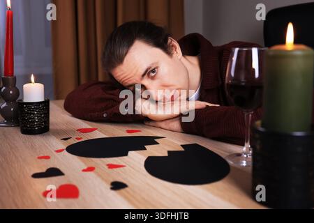 Deprimierter Mann, der auf seinen Armen an einem Tisch liegt, mit gebrochenem schwarzem Herzen und Wein. Trauriger einsamer Kerl, der unter Trennung leidet. Anti-Valentinstag-Konzept. Hallo Stockfoto