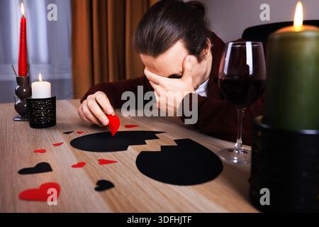 Ein weinender Mann mit Gesicht in der Hand sitzt am Tisch mit Wein und gebrochenem schwarzem Herzen. Trauriger einsamer Kerl, der unter Trennung leidet. Anti-Valentinstag-Konzept. H Stockfoto