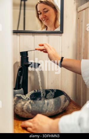 Junge kaukasische Frau mit langen Haaren wäscht sich die Hände an einem stilvollen Steinbecken in einer gemütlichen Ferienhütte. Ein Spiegel spiegelt ihren fokussierten Ausdruck wider. Stockfoto