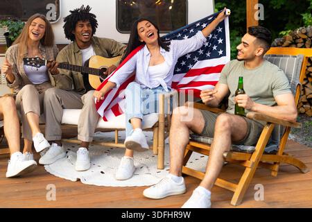 Multinationale Freunde spielen Gitarre und singen, trinken Bier, halten amerikanische Flagge, sitzen auf einem Campingausflug in der Nähe von Wohnmobil Stockfoto