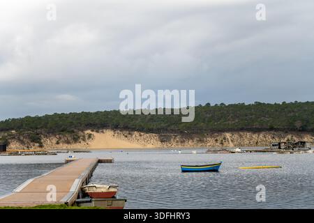 Sesimbra Portugal 25. Dezember 2025. Holzpier und kleine Boote sitzen auf Lagunenwasser mit Sanddünen und Wald im Hintergrund Stockfoto