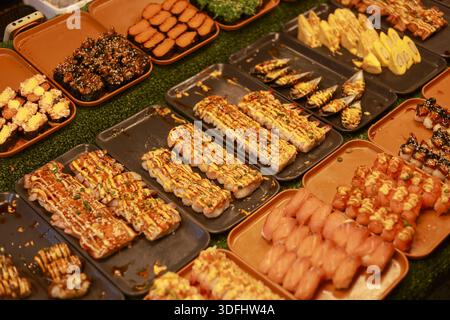 Köstliche Auswahl an Sushi-Brötchen und Nigiri, die auf Tabletts am pulsierenden Nachtmarktstand mit appetitlichen Belägen wie Krabbenstock, Lachs und Süßigkeiten ausgestellt werden Stockfoto