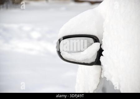 Eine Nahaufnahme des komplett mit dichtem Schnee bedeckten Seitenspiegels eines Autos mit einer verschwommenen Winterszene Stockfoto