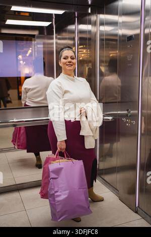 Fröhlicher Shopper mit lebendigen Taschen in einer modernen Fahrstuhlumgebung Stockfoto