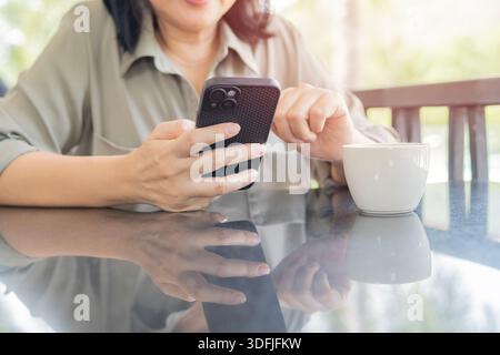 Abgeschnittene Aufnahme einer Frau in einem grünen Hemd mit einem Handy in der Nähe des Balkons im Landhaus mit einer Tasse Kaffee. Stockfoto