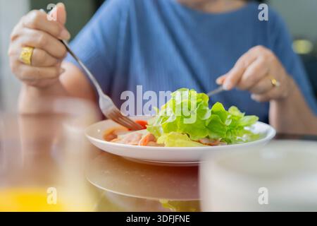 Eine ältere Frau, die im Speisesaal frühstückt und Gemüse- und Specksalat isst und Kaffee trinkt. Selektiver Fokus. Stockfoto