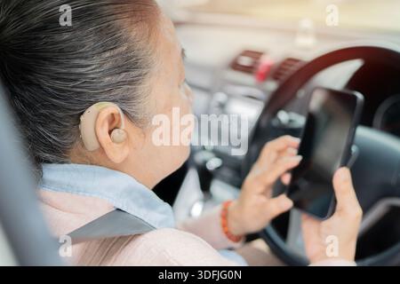 Asiatische Großmutter mit Hörgerät, die Handy mit Hilfe der Assistentin-App künstlicher Intelligenz im Auto hält. Selektiver Fokus. Stockfoto