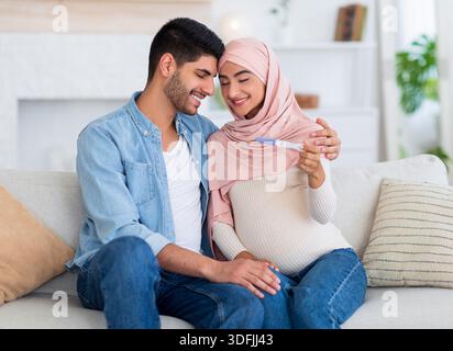 Familie und Beziehungskonzept. Schwangere muslimische Paare mit Schwangerschaftstest zu Hause lieben, umarmen und lächeln Stockfoto