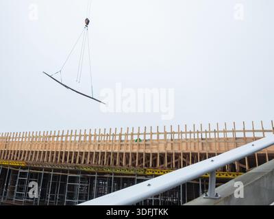 Eine Bauszene mit Holzschalung und Metallgerüsten, wobei ein Kran einen gebogenen Metallbalken gegen einen bewölkten Himmel senkt Stockfoto