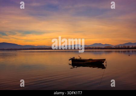 Kerkini, Nordgriechenland – Ein Holzboot schwimmt auf ruhigen Gewässern bei Sonnenuntergang und reflektiert warme Himmelsfarben in einer ruhigen und friedlichen Landschaft. Stockfoto