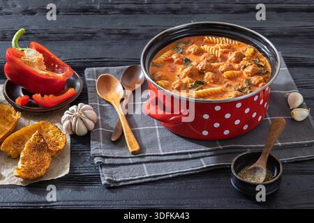 Italienische Fusilli-Nudelsuppe mit Fleischbällchen und Spinat in cremiger Tomatensoße in rotem Topf auf schwarzem Holztisch mit geröstetem Brot, Knoblauch und Löffeln, Hor Stockfoto
