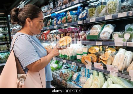 Eine Frau steht in einem Supermarkt und sieht sich einen Kürbis genau an. Sie hält es in einer Hand, während sie verschiedene verpackte Gemüsesorten in den Regalen beobachtet Stockfoto