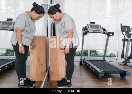 Eine Frau steht vor einem Spiegel in einem Fitnessstudio und konzentriert sich auf ihr Spiegelbild. Sie trägt Trainingskleidung und ist neben Trainingsgeräten. Das Zimmer Stockfoto