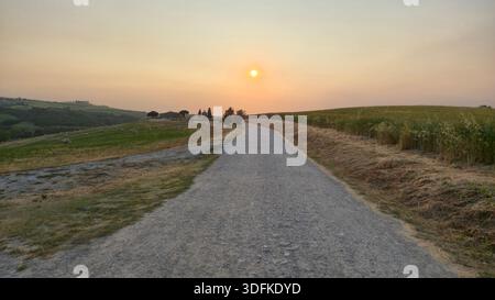 Gravel Road, die in Richtung einer traditionellen Steinkapelle (Capella madonna di vitalete) in den Rolling Hills der Toskana bei Sonnenuntergang der Golden Hour führt Stockfoto