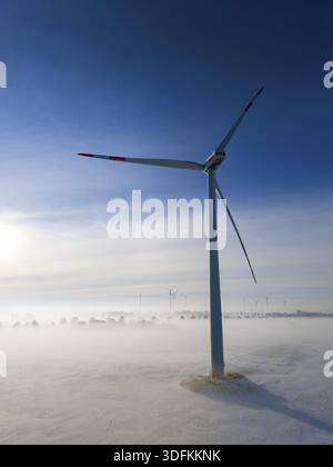 Eine Windturbine hebt sich in einer schneebedeckten, nebeligen Landschaft hervor, umgeben von sanftem Morgenlicht und klarem Himmel, Luftsicht, Windturbine im Schnee im Nebel, zwischen Stockfoto