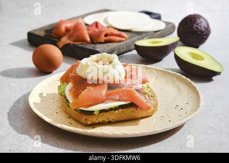 Toast mit Lachs, pochiertem Ei, Ziegenkäse und Avocado. In Scheiben geschnittene Zutaten wie Lachs und Käse befinden sich auf einem Holzbrett. Ein Ei ist in der Nähe Stockfoto
