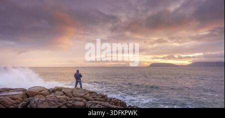Fotograf fotografiert Wellen, die auf Felsen stürzen, mit erstaunlichem Sonnenaufgang im Hintergrund. Fanad Head Peninsula am Wild Atlantic Way, Donegal, Irland Stockfoto