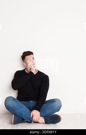Junger Mann in lässiger Kleidung, der an einem ruhigen Nachmittag nachdenklich an einer Weißen Mauer sitzt Stockfoto