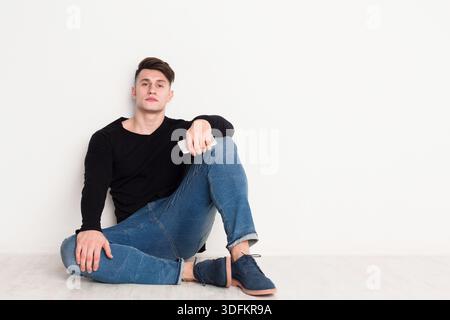 Junger Mann, der lässig an einer weißen Wand sitzt und ein Smartphone in entspannter Pose hält Stockfoto