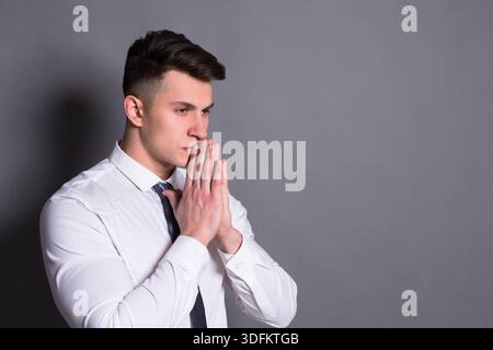 Mann in einem weißen Hemd und Krawatte, der nachdenklich vor einem grauen Hintergrund nachdenkt Stockfoto