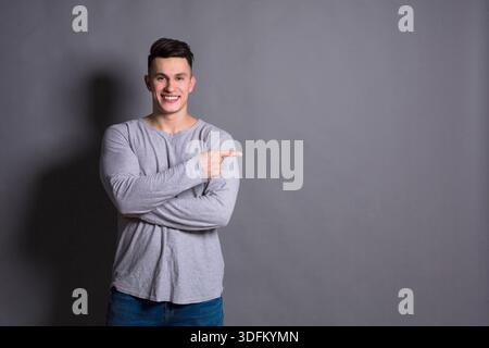 Junger Mann posiert lächelnd und zeigt zur Seite vor grauem Hintergrund Stockfoto