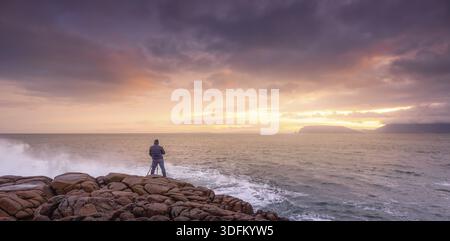 Fotograf fotografiert Wellen, die auf Felsen stürzen, mit erstaunlichem Sonnenaufgang im Hintergrund. Fanad Head Peninsula am Wild Atlantic Way, Donegal, Irland Stockfoto