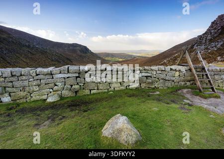 Trauernde Mauer mit Leiter auf der Hare-Lücke, die das schöne Tal mit blauem Himmel und weißen Wolken beaufsichtigt. Mourne Mountains Range in Nordirland Stockfoto